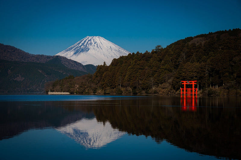 Snow-capped Mt. Fuji rising behind forested hills on Lake Ashi, with Hakone Shrine’s red torii standing in the water and reflected in the still lake.