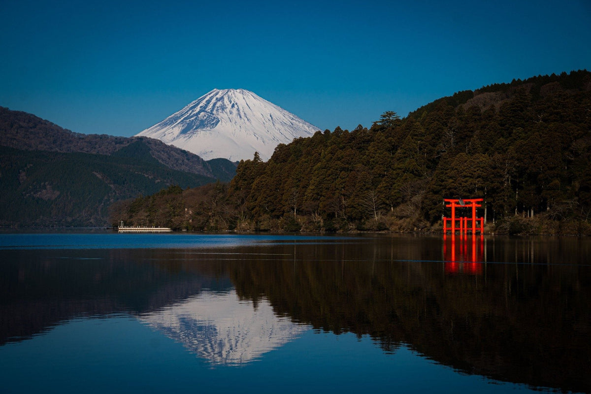 Snow-capped Mt. Fuji rising behind forested hills on Lake Ashi, with Hakone Shrine’s red torii standing in the water and reflected in the still lake.