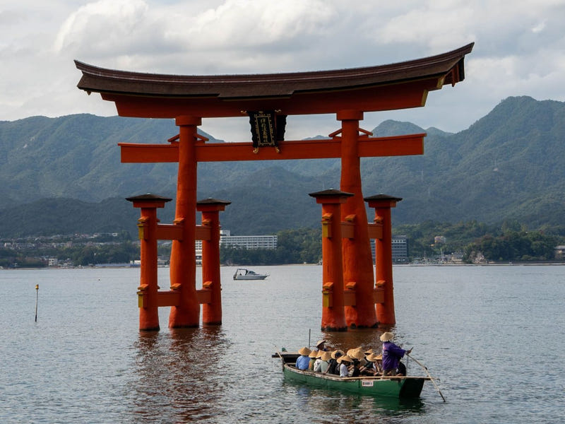 Itsukushima Shrine’s vermilion great torii standing in the sea at high tide, with a wooden rowing boat of visitors in sedge hats passing in front and green mountains behind.
