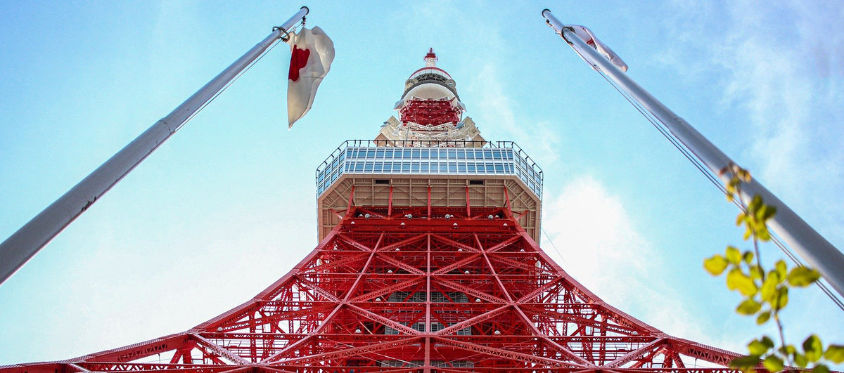 Looking straight up Tokyo Tower’s red steel lattice to the main deck and spire, framed by two flagpoles and a Japanese flag against a blue sky.