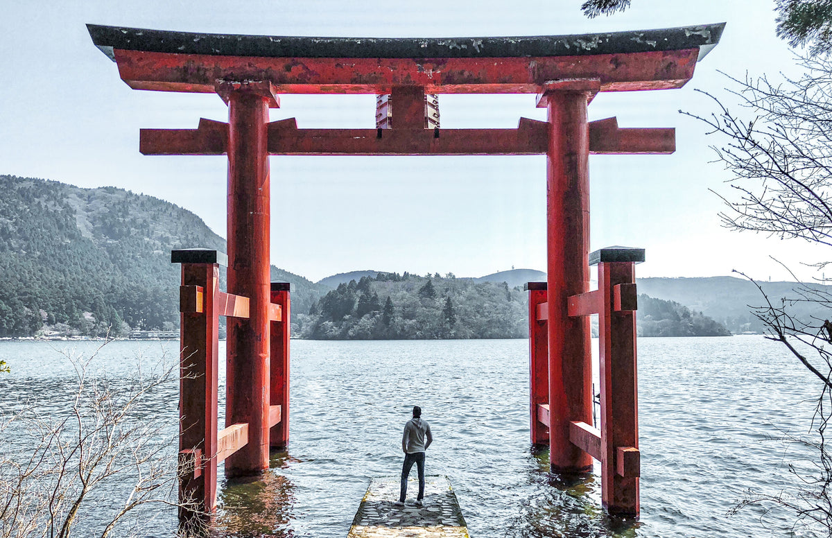 A lone figure on a stone pier beneath a red torii gate standing in a lake, wooded hills behind.