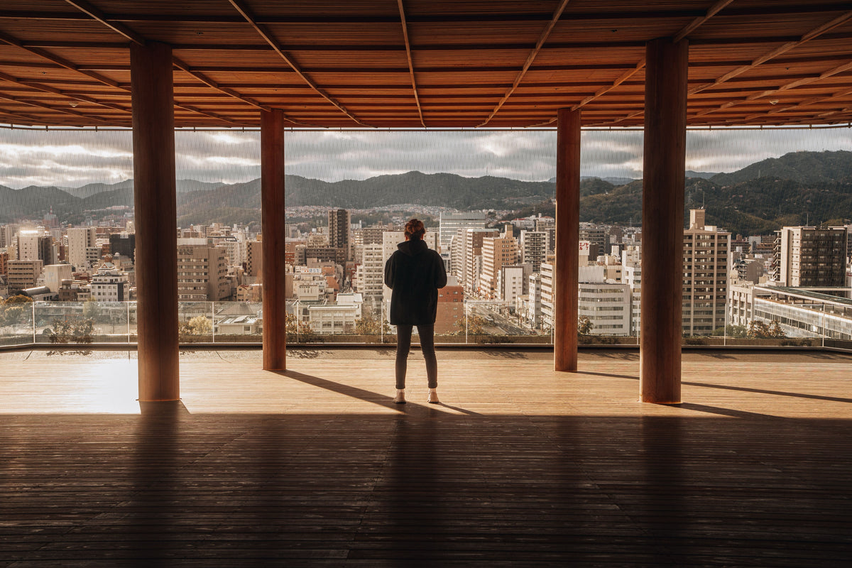 A figure on a wooden viewing deck looking out over a Japanese city and the hills behind it.