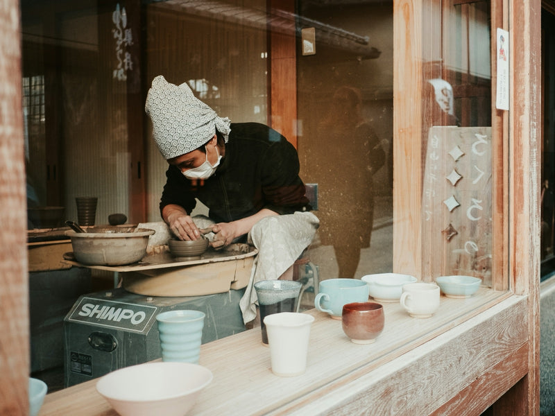 A potter shaping clay at a wheel inside a workshop, with finished cups on the window sill.