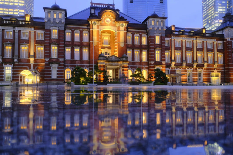 The brick facade of Tokyo Station reflected in the glass of a modern tower.